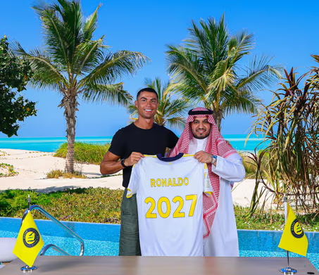 Cristiano Ronaldo with an Arabian man holding Ronaldo 2027 jersey while he pens new contract at Al Nassr